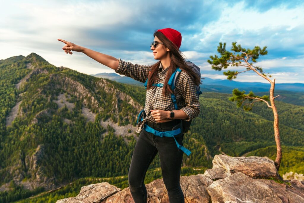 A young tourist pointing her finger at the mountains. A tourist in sunglasses and a backpack walking along the rocks in the mountains, smiles and points to something in front of her. Travel and hiking